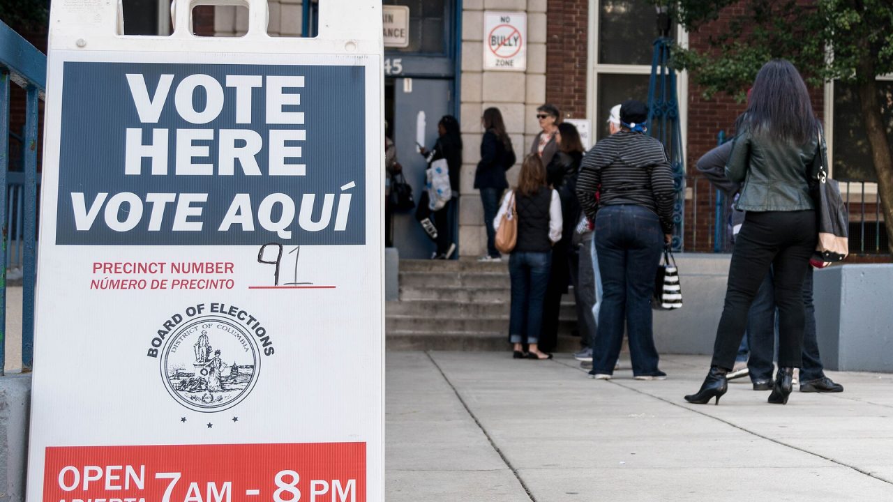 Voters wait outside a polling place next to a bilingual "Vote Here / Vote Aquí" sign.